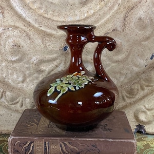May include: A brown ceramic vase with a green floral design and a handle. The vase is sitting on top of two books.