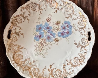 May include: A white porcelain serving plate with a gold floral design and blue flowers. The plate has two handles on the sides.