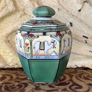 May include: A decorative ceramic jar with a lid. The jar has a green base and lid, with a cream-colored body featuring a camel and Egyptian-style illustrations. The jar sits on a patterned surface.