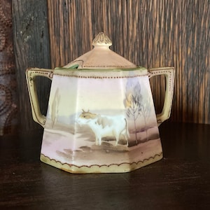 May include: A white porcelain sugar bowl with a lid and two handles. The bowl is decorated with a painted scene of a cow in a field. The bowl has a light pink, green, and brown color scheme.