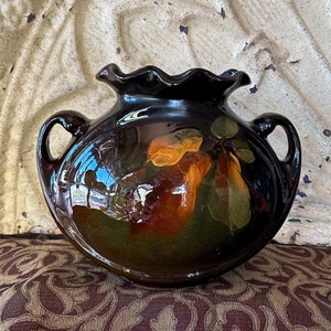 May include: A brown ceramic vase with two handles and a scalloped rim. The vase is decorated with a painted design of a brown, orange, and yellow fruit with leaves.