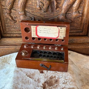 May include: A wooden box containing a set of brass weights and a pair of tweezers. The lid features a white label with handwritten text including 20-.002gm, 10-.0012gm, and 5-.0008gm. The interior has circular cutouts for weights and a black insert.