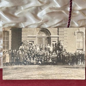 May include: A vintage black and white photograph depicting a large group of people posed in front of a brick building with arched windows and a central doorway. The individuals are arranged in multiple rows on steps, wearing early 20th-century attire.