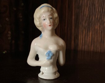 May include: A white porcelain doll with a blue ribbon and a blue flower in her hand. The doll is a vintage pincushion.