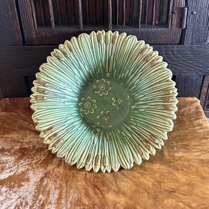 Puede incluir: Un cuenco de cerámica verde con un diseño de girasol. El cuenco tiene un borde acanalado y un centro con tres pequeños diseños de flores. El cuenco está sobre una superficie marrón.