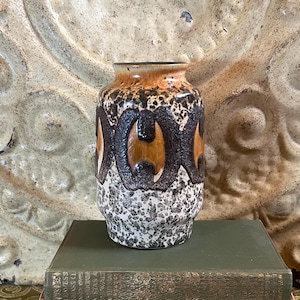 May include: A ceramic vase featuring a cylindrical shape with a textured surface. The upper portion is a smooth orange glaze, while the middle band displays dark brown, raised geometric patterns. The base has a speckled white and gray finish.