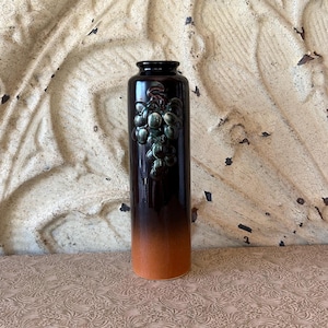 May include: A tall, cylindrical vase with a dark brown to orange gradient. A cluster of green grapes is embossed on the front. The vase is likely ceramic or stoneware, and it has a glossy finish. The vase is a decorative item.