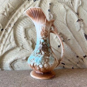 Puede incluir: Jarra de cerámica vintage con asa, con parte superior acampanada y cuerpo bulboso. La jarra está adornada con patrones florales marrones, blancos y turquesas. Tiene una base de pedestal y un asa curva.