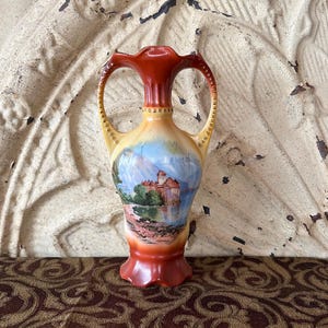 May include: A decorative ceramic vase with a landscape painting. The vase has a flared rim and two handles. The colors include shades of orange, yellow, and blue. The vase is approximately 7 inches tall.