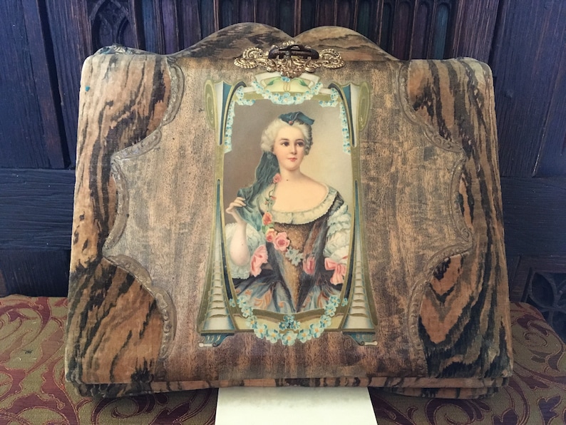 Puede incluir: Un &aacute;lbum de fotos vintage con una cubierta de madera y un dise&ntilde;o floral decorativo. La cubierta presenta un retrato de una mujer con un vestido azul y blanco con un ramo de flores.