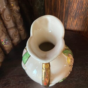 Puede incluir: Una jarra de cer&aacute;mica blanca con asa dorada y dise&ntilde;o floral. La jarra tiene flores y hojas rosas y verdes.
