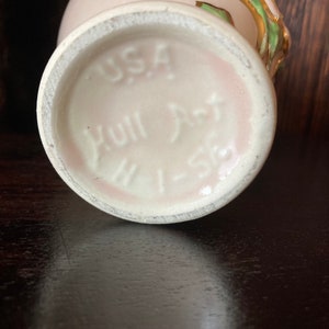 Puede incluir: Un jarr&oacute;n de cer&aacute;mica blanca con un esmalte rosa y detalles dorados. El jarr&oacute;n tiene un dise&ntilde;o floral con hojas verdes y una flor rosa. La base del jarr&oacute;n est&aacute; marcada con "USA Hull Art 1-58".