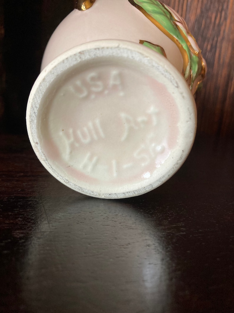 Puede incluir: Un jarr&oacute;n de cer&aacute;mica blanca con un esmalte rosa y detalles dorados. El jarr&oacute;n tiene un dise&ntilde;o floral con hojas verdes y una flor rosa. La base del jarr&oacute;n est&aacute; marcada con "USA Hull Art 1-58".