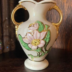 Puede incluir: Un peque&ntilde;o jarr&oacute;n de cer&aacute;mica blanca con detalles dorados y un dise&ntilde;o floral. El jarr&oacute;n tiene dos asas y un borde festoneado.