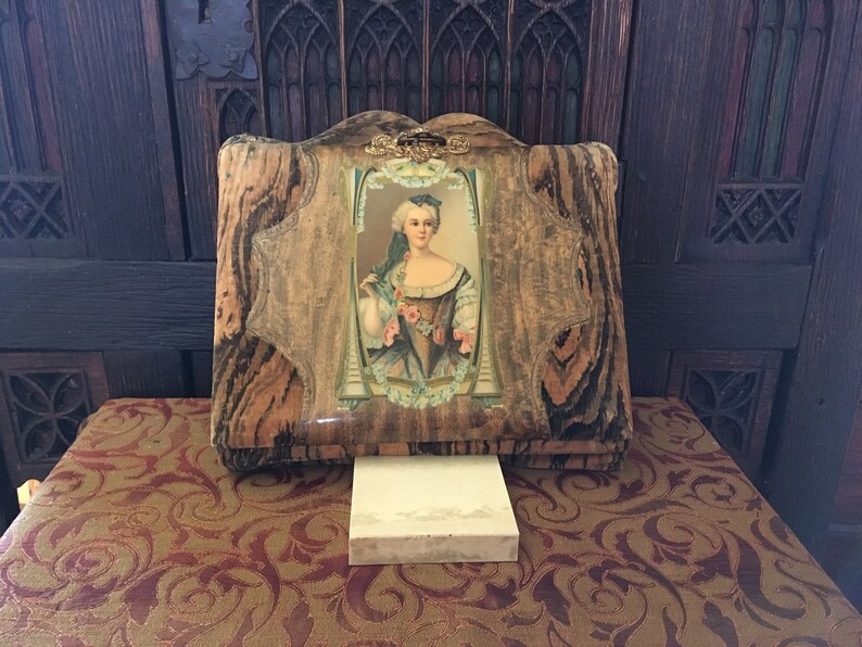 May include: A closed, antique photo album with a wood-grain cover. The cover has a decorative gold metal clasp and a small picture of a woman in a blue dress with a floral pattern. The album is sitting on a brown surface with a floral pattern.