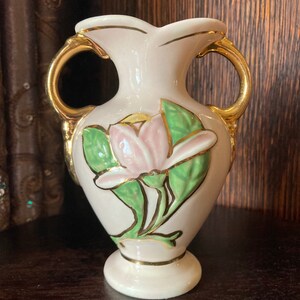 Puede incluir: Un peque&ntilde;o jarr&oacute;n de cer&aacute;mica blanca con detalles dorados y un dise&ntilde;o floral rosa y verde. El jarr&oacute;n tiene dos asas y una base redonda.