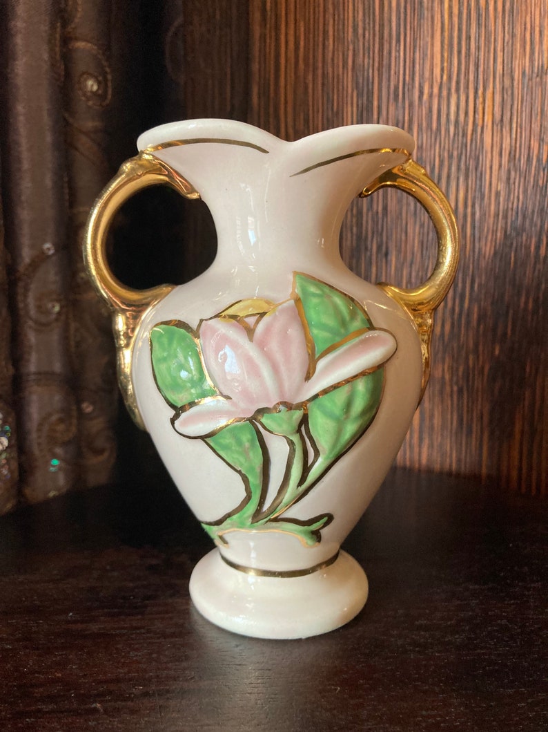 Puede incluir: Un peque&ntilde;o jarr&oacute;n de cer&aacute;mica blanca con detalles dorados y un dise&ntilde;o floral rosa y verde. El jarr&oacute;n tiene dos asas y una base redonda.