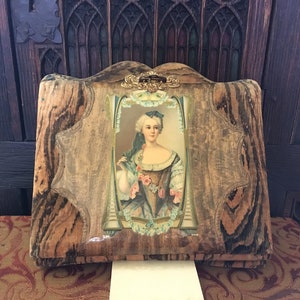 Puede incluir: Un &aacute;lbum de fotos vintage de madera con un dise&ntilde;o decorativo y ornamentado. La portada presenta una imagen enmarcada de una mujer con un vestido azul y blanco con estampado floral. El &aacute;lbum tiene un cierre de lat&oacute;n.