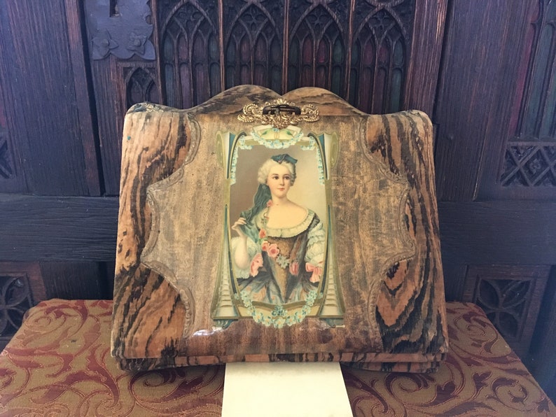 Puede incluir: Un &aacute;lbum de fotos vintage de madera con un dise&ntilde;o decorativo y ornamentado. La portada presenta una imagen enmarcada de una mujer con un vestido azul y blanco con estampado floral. El &aacute;lbum tiene un cierre de lat&oacute;n.