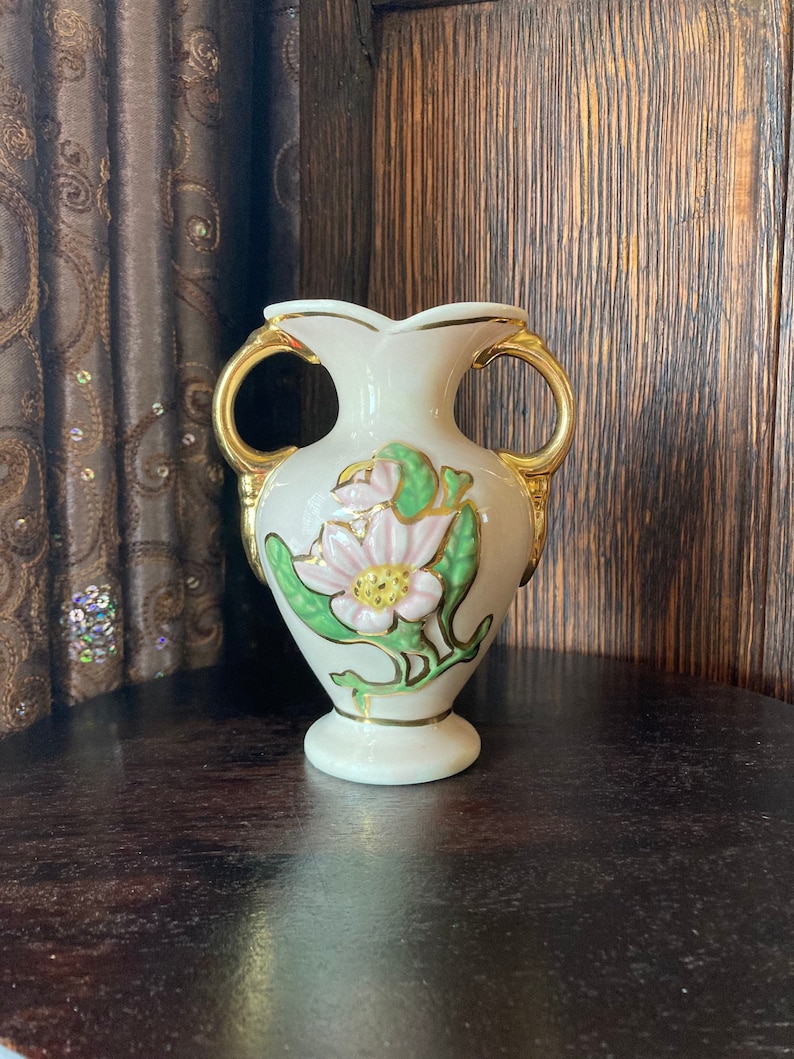 Puede incluir: Un peque&ntilde;o jarr&oacute;n de cer&aacute;mica blanca con detalles dorados y un dise&ntilde;o floral rosa y verde. El jarr&oacute;n tiene dos asas y una base redonda.