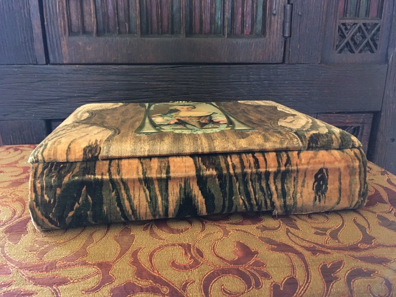Puede incluir: Una caja vintage cubierta de terciopelo con un patr&oacute;n de veta de madera y un dise&ntilde;o floral en la parte superior. La caja est&aacute; cerrada y se encuentra sobre una superficie estampada.