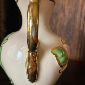 Puede incluir: Un jarr&oacute;n de cer&aacute;mica blanca con un asa dorada y un dise&ntilde;o floral. El jarr&oacute;n tiene un patr&oacute;n floral verde y rosa con detalles dorados.