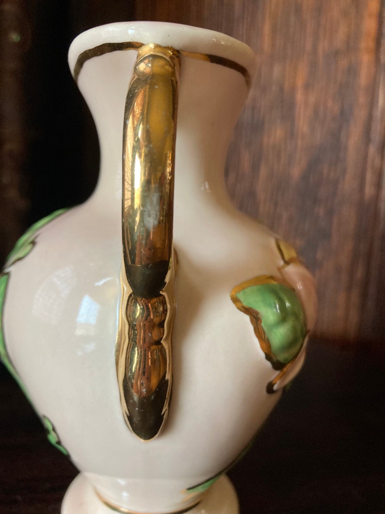 Puede incluir: Un jarr&oacute;n de cer&aacute;mica blanca con un asa dorada y un dise&ntilde;o floral. El jarr&oacute;n tiene un patr&oacute;n floral verde y rosa con detalles dorados.