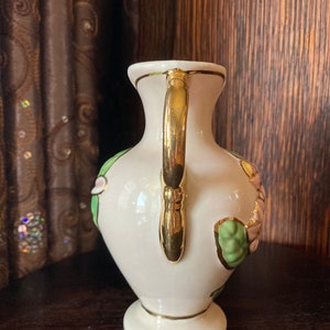 Puede incluir: Un jarr&oacute;n de cer&aacute;mica blanca con un asa dorada y detalles florales. El jarr&oacute;n tiene un dise&ntilde;o floral verde y rosa con acentos dorados.