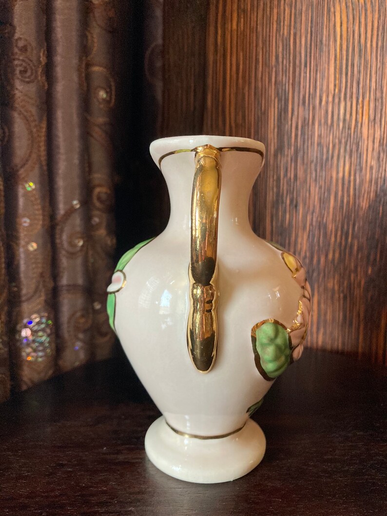 Puede incluir: Un jarr&oacute;n de cer&aacute;mica blanca con un asa dorada y detalles florales. El jarr&oacute;n tiene un dise&ntilde;o floral verde y rosa con acentos dorados.