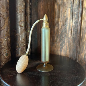 Può includere: Un vaporizzatore di profumo vintage in vetro con un top dorato e un atomizzatore a bulbo. Il vaporizzatore è su un tavolo di legno con una superficie marrone.