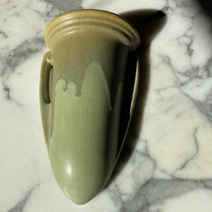 Puede incluir: Un jarrón de pared de cerámica verde salvia con un esmalte craquelado. El jarrón tiene una parte superior redondeada con dos asas pequeñas y una base puntiaguda. El jarrón mide aproximadamente 20 cm de alto.