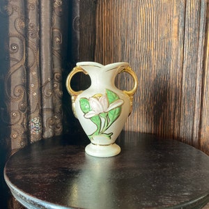 Puede incluir: Un jarr&oacute;n de cer&aacute;mica blanco con detalles dorados y un dise&ntilde;o floral. El jarr&oacute;n tiene dos asas y un delicado patr&oacute;n floral con hojas verdes y una flor rosa.