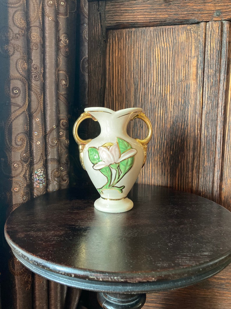 Puede incluir: Un jarr&oacute;n de cer&aacute;mica blanco con detalles dorados y un dise&ntilde;o floral. El jarr&oacute;n tiene dos asas y un delicado patr&oacute;n floral con hojas verdes y una flor rosa.