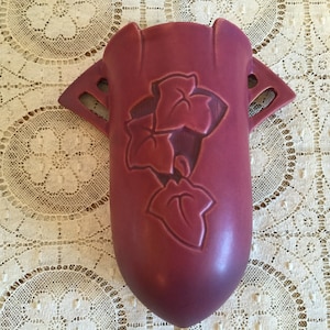 May include: A deep burgundy ceramic wall vase with a floral design. The vase has two handles and a rounded bottom.