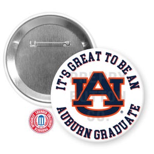 May include: White button with the text "IT'S GREAT TO BE AN AUBURN GRADUATE" in navy blue and orange. The button features the Auburn University logo. A second button is visible, showing the pin back. A licensed collegiate products seal is also visible.