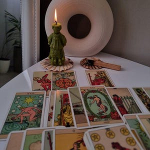 Puede incluir: Una vela verde encendida con forma de figura se encuentra sobre una concha, rodeada de cartas del tarot. Un jarrón blanco con una abertura circular está en el fondo. Las cartas son coloridas con varias ilustraciones.