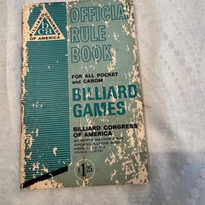 May include: A vintage billiard rule book with a teal and white cover. The cover features the text "OFFICIAL RULE BOOK" and "BILLIARD GAMES". The book is from the Billiard Congress of America.