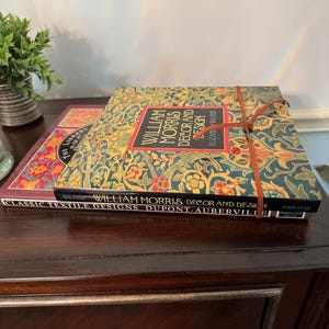 May include: Two hardcover books stacked on each other. The top book is titled William Morris Decor and Design by Elizabeth Wilhide, featuring a teal, gold, and red floral pattern and a brown leather tie. The bottom book is Classic Textile Designs.