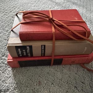 Könnte beinhalten: Ein Stapel von drei alten Büchern, die mit einem braunen Lederriemen zusammengebunden sind. Die Bücher haben rote und beige Einbände, wobei das mittlere Buch den Titel "The Complete Essays of Mark Twain" trägt.