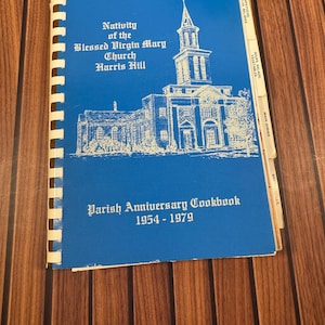May include: A blue spiral-bound cookbook titled Nativity of the Blessed Virgin Mary Church Harris Hill Parish Anniversary Cookbook 1954-1979. The cover features a white line drawing of a church and includes side tabs for recipe categories.