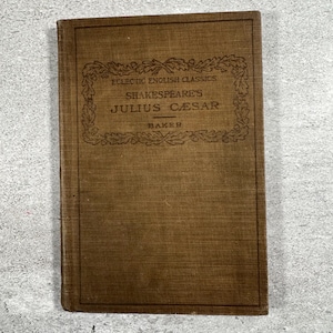 May include: A brown hardcover book titled Eclectic English Classics Shakespeare's Julius Caesar edited by Baker. The front cover features a dark brown rectangular border with an oak leaf pattern and centered black text on the fabric surface.