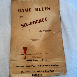 Puede incluir: Libro de reglas de juego vintage para billar Six-Pocket. La portada es de color beige con texto rojo que dice "GAME RULES" y "SIX-POCKET". El folleto también enumera "Reglas generales", "Faltas" y otras variaciones del juego de billar. El fabricante es Fischer Manufacturing Co., Inc.