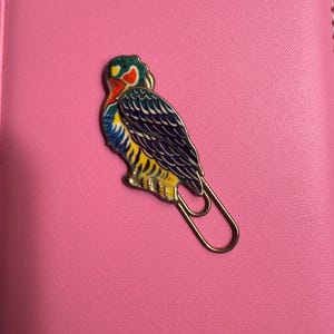 May include: A colorful bird-shaped paperclip with a gold-tone finish. The bird features a detailed design with red, blue, yellow, and green enamel. The paperclip is resting on a pink surface.