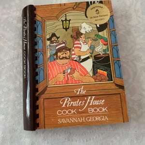 May include: A spiral-bound cookbook titled The Pirate's House COOK BOOK SAVANNAH, GEORGIA. The cover features a cartoon illustration of pirates in a kitchen, a hanging lantern, and a gold seal stating Improved 38 New Recipes REVISED.