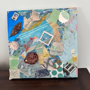 May include: Mixed media collage on canvas with various textures and colors. Features a quote, a clock face, a small framed picture, and a brown leather object. The background includes map fragments and a blue and white color scheme.