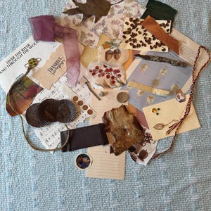 Può includere: Una collezione di materiali artistici misti, tra cui carta, nastro ed elementi naturali come foglie e pezzi di legno. Gli oggetti sono disposti su una superficie trapuntata azzurra, creando un'estetica rustica e artistica.