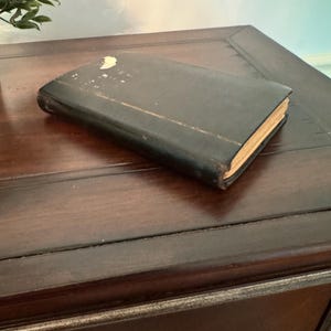 May include: A rectangular book with a worn black cover featuring a small area of white damage on the top edge and visible light-colored pages along the side. The book is positioned at an angle, showing its front cover and the thickness of the pages.