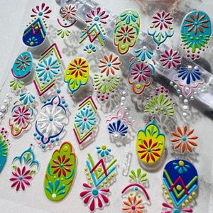 Puede incluir: Una hoja de pegatinas adhesivas para uñas con una variedad de coloridos patrones florales y geométricos en relieve en tonos azul, verde, rosa, naranja y blanco. Los diseños incluyen diamantes, pétalos y motivos circulares con el texto JP24-1028.