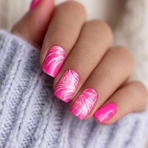 May include: Close-up of a hand with manicured nails. The nails are painted a vibrant pink with a white palm leaf design. The hand is resting on a light blue knitted sweater, creating a contrast of colors and textures.