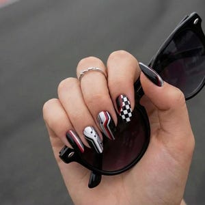 May include: A hand holding black sunglasses with oval lenses. The nails are painted with a variety of designs, including a checkered pattern, red and white stripes, and abstract shapes. A silver ring is on the ring finger.